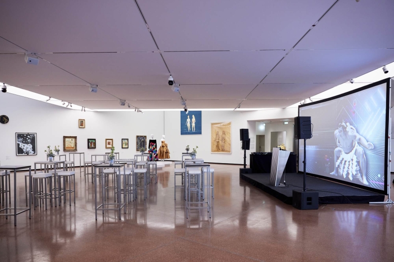 Moderne Galerie mit Tischen, Kunstwerken an Wänden und einer Präsentationsleinwand.