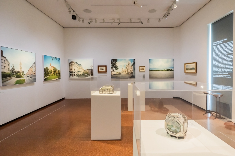 Das Foto zeigt einen Ausstellungsraum mit vielen Bildern an den Wänden und drei Vitrinen in der Mitte. Zu sehen ist ein Querschnitt aus historischen und zeitgenössischen Stadtansichten von Wien.
An den Wänden hängen großformatige Fotografien von Stefan Oláh, die Wiener Plätze, Straßen und Gebäude in ruhiger, klarer Bildsprache zeigen. Dazwischen sind Aquarelle von Rudolf von Alt.
In den Vitrinen stehen Dosen mit Wienansichten von Balthasar Wigand. Jede Dose zeigt mehrere Ansichten – zum Beispiel den Stephansdom oder die Donau.
Rechts an der Wand befindet sich ein Ausstellungstext.