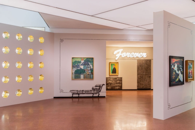 The photo shows part of the “Klimt⇄Warhol” exhibition in the Heidi Horten Collection museum in Vienna.
On the left you can see a wall with round windows that look like portholes.
This wall was designed by the artist Markus Schinwald.
In the middle, directly in front of us, hangs a painting by Gustav Klimt.
It shows the Attersee, a lake in Austria.
The picture shows water, houses and nature in bright colors.
Markus Schinwald also designed the decorations on the walls and the painted pieces of furniture next to and under the works of art.
At the back of the wall, above a picture, the word “Forever” is illuminated in large white letters.
This illuminated lettering is a work of art by Tim Noble & Sue Webster.
Below it hangs a self-portrait by Edvard Munch.
On the far right, another wall juts diagonally into the room.
Two paintings by Emil Nolde, which are also part of the collection, hang there.