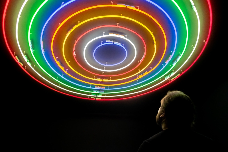 Neonringe in verschiedenen Farben leuchten an der Decke, während eine Person nach oben schaut. Im Hintergrund ein weiteres Kunstwerk.