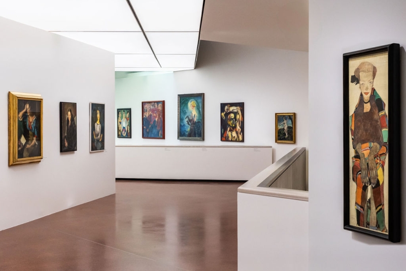 The photo shows a bright room in the exhibition “Experiment Expressionism.”
There are many paintings hanging on the white walls.
The pictures show women and men – some look calm, others are moving or appear strong.
On the far right, you see a long, narrow painting of a woman.
The painting is by Egon Schiele.
On the left wall, there are three portraits hanging next to each other.
In the middle of the room, you see more paintings, for example the picture “Terzetta”, with the woman and the long hands.
The floor is brown, and the ceiling is bright.