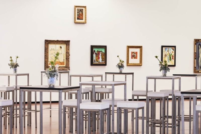 Moderne Galerie mit hohen Tischen, weißen Stühlen und mehreren gerahmten Kunstwerken an der Wand.
