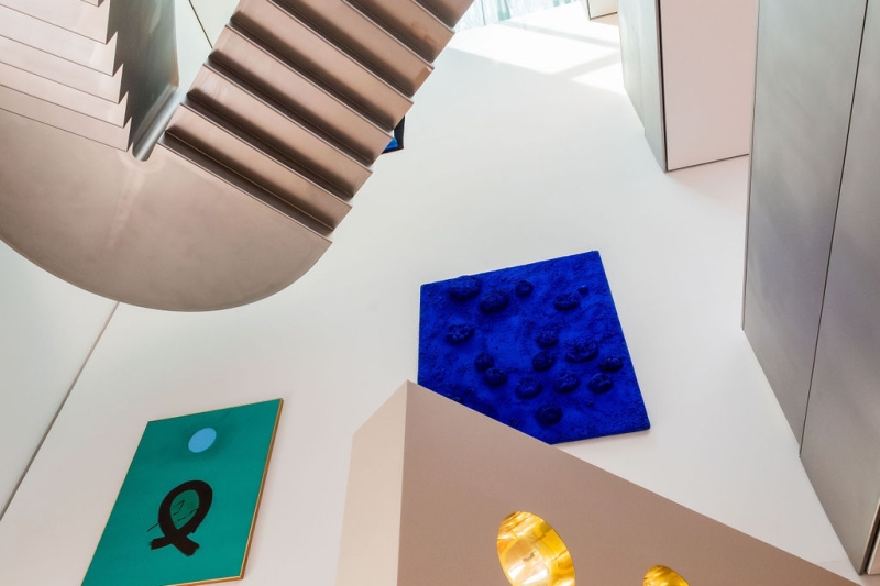 The photo shows a view upstairs in the Heidi Horten Collection museum in Vienna.
You can see a modern metal staircase and large, bright walls.
On the left wall hangs a green picture with a black sign and a blue circle.
This work of art is called “Blue Orb” and is by Adolph Gottlieb.
Next to it, further up, you can see a bright blue work of art with sponges on it.
This is the very first sponge relief by Yves Klein.
He has dyed real sponges with deep blue paint and glued them onto the canvas.
The work is very special because Yves Klein became famous for this intense blue color.
At the bottom of the picture you can see part of Markus Schinwald's porthole wall.
The round, shiny gold windows are part of a design element.