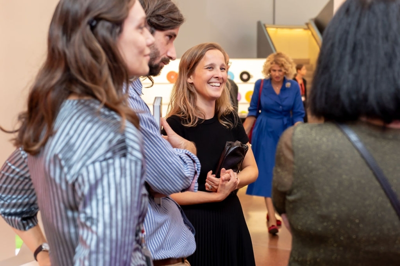 Auf dem Bild sieht man eine Gruppe von Menschen in der Heidi Horten Collection.
Alle schauen sich etwas an und unterhalten sich.
In der Mitte des Fotos steht eine Frau mit blondem Haar.
Sie trägt ein schwarzes Kleid und lächelt freundlich.
Um sie herum stehen mehrere Personen – sie sehen interessiert und entspannt aus.
Im Hintergrund sieht man eine Frau im blauen Kleid, die ebenfalls durch den Raum geht.
Die Stimmung auf dem Foto wirkt offen, freundlich und neugierig