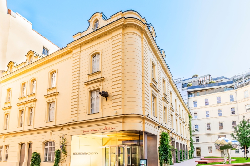 Elegantes Gebäude mit gelber Fassade, Glas-Eingang und grüner Bepflanzung, umgeben von modernen Strukturen.