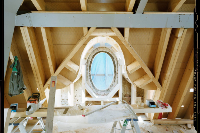 This picture shows the interior of a roof truss during construction work. The focus is on a large, oval window surrounded by a wooden frame. The walls and roof slopes are made of light-colored wood, which gives the room a warm atmosphere - despite the construction site situation. There are ladders and tools on the floor and a spirit level leaning against a beam. The blue protective fleece in front of the window lets soft light into the room.