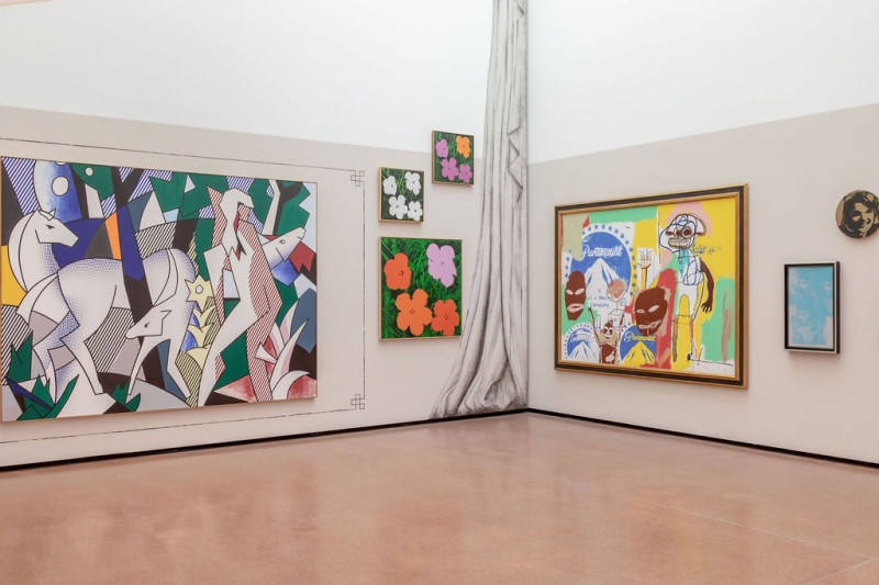 The photo shows part of the "Klimt⇄Warhol" exhibition in the Heidi Horten Collection museum in Vienna.
The photo shows a corner of the exhibition space,
where two walls meet. Several colorful paintings hang on these walls.
On the far left is a large painting by Roy Lichtenstein.
It shows several white deer in an abstract landscape,
with strong colors and black outlines - typical of Lichtenstein's style, which is reminiscent of comics.
To the right, hung one above the other, are three small pictures of colorful flowers.
These floral prints are by Andy Warhol. They are very colorful, with simple shapes and strong contrasts.
In the right-hand corner hangs a large, joint painting by Jean-Michel Basquiat and Andy Warhol.
The picture is wild, colorful and full of energy.
You can make out figures, writing and faces.
To the right is another print by Andy Warhol -
a small, light blue picture showing Elisabeth Taylor as Cleopatra.
Above it, round in gold, is another work by Warhol:
a portrait of Jackie Kennedy, the former First Lady of the USA.
A painted gray curtain visually separates the two walls and is reminiscent of a theater or stage.
This part of the wall design is also part of Markus Schinwald's staged exhibition design.