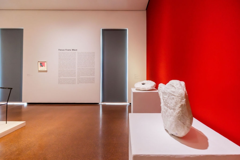 Art gallery interior featuring a red wall, sculptures on pedestals, and an informational text panel.