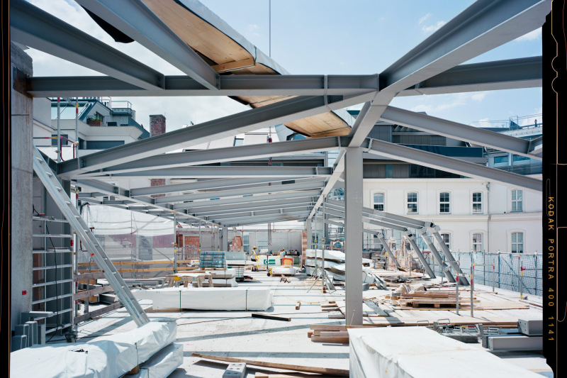 The photo shows a construction site with an impressive steel structure. The massive girders span the space and give a first impression of the future architecture. Building materials, tools and equipment are neatly arranged all around. A bright existing building can be seen in the background, forming an exciting contrast to the modern steel structure. The sky is blue, the light bright - a snapshot of the construction process in progress. The photo was taken by photographer Stefan Oláh.