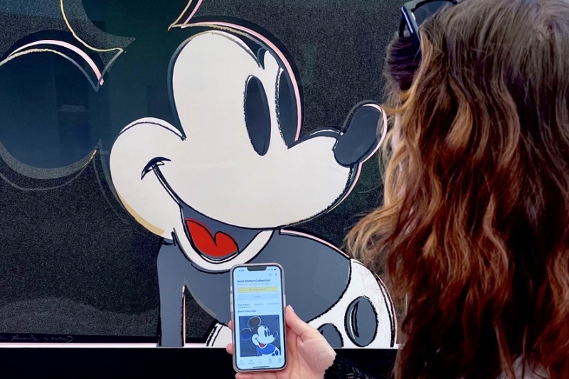 Auf dem Foto sieht man eine Frau von hinten, die sich ein Kunstwerk von Andy Warhol anschaut. Auf dem Bild ist Micky Maus. Die Frau hält ein Smartphone in der Hand. Darauf ist die App Smartify geöffnet – ein Audioguide, der Informationen über Kunstwerke gibt. Man sieht, dass sie genau das Bild auf dem Handy geöffnet hat, das auch vor ihr an der Wand hängt. Das Foto zeigt, wie digitale Medien im Museum helfen, mehr über Kunst zu erfahren – einfach und direkt am eigenen Handy
