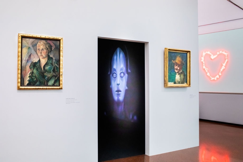 The photo shows a doorway in the exhibition “Experiment Expressionism. Schiele meets Nosferatu”.
In the room behind the doorway, there is a large glowing image of the machine figure from the film “Metropolis” (1927).
On the left and right sides of the doorway, there are two portraits in golden frames:
On the left is a sharp-edged, serious portrait of a woman.
On the right is a smaller, more loosely painted portrait with a blue-green background and a yellow hat – this is the self-portrait of Herbert Boeckl.
In the background on the right, a pink neon heart is glowing on the wall.