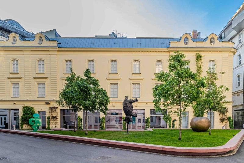 The photo shows the Heidi Horten Collection building in Vienna.
It is a bright yellow house with a neoclassical façade and large windows.
The building is located in the Hanuschhof, a quiet courtyard in the middle of the city.
In front of the museum is a small sculpture garden with grass, trees and works of art.
You can see three different sculptures there:
On the far left is a green figure by Keith Haring.
In the middle is a dark bronze figure, an elephant by Miquel Barceló.
On the far right is a large golden apple. It is by Claude Lalanne.