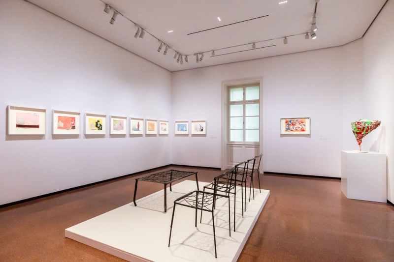 Art gallery interior featuring framed artworks on the wall, a modern table, and metal chairs on a raised platform.
