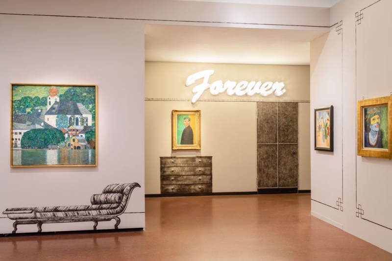 The photo shows part of the "Klimt⇄Warhol" exhibition in the Heidi Horten Collection museum in Vienna.
On the left, hangs a painting by Gustav Klimt.
It shows Lake Attersee, a lake in Austria.
The painting shows water, houses and nature in bright colors.
Markus Schinwald also designed the decorations on the walls and the painted pieces of furniture next to and under the works of art.
At the back of the wall, above a picture, the word “Forever” is illuminated in large white letters.
This illuminated lettering is a work of art by Tim Noble & Sue Webster.
Below it hangs a self-portrait by Edvard Munch.