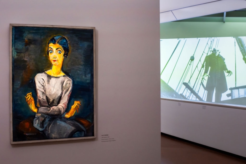 The photo shows a part of the exhibition “Experiment Expressionism.”
On the left, there is a painting by Lilly Steiner.
It shows Lilian Gärtner with big blue eyes and a grey dress.
She is sitting and holding her hands in a special, expressive way.
This painting was already described earlier.
On the right, you see a film scene from “Nosferatu.”
The film is black and white and is projected onto the wall.
You can see the vampire Nosferatu standing on a ship.
He is on the deck and looks scary.