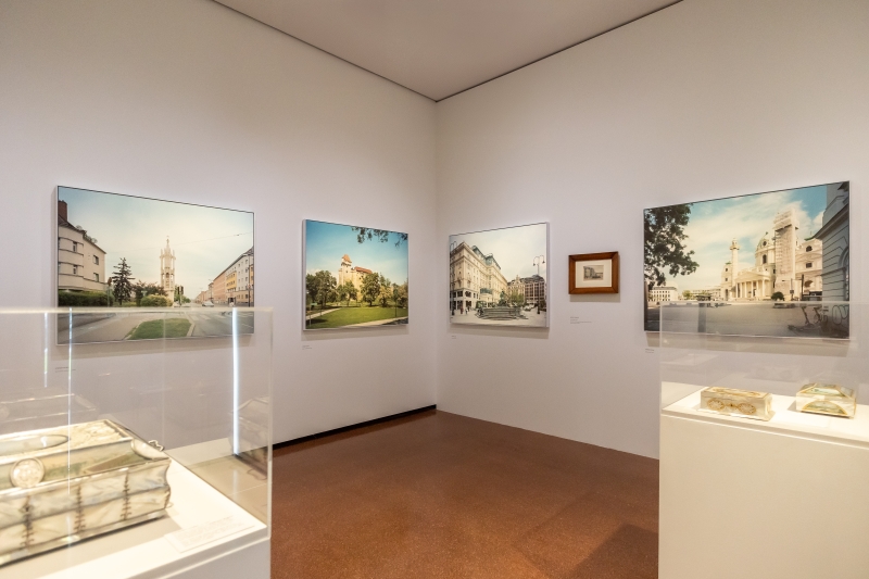 Das Foto zeigt einen Ausstellungsraum mit mehreren Bildern an den Wänden und zwei Vitrinen in der Mitte. Zu sehen ist ein Querschnitt aus historischen und zeitgenössischen Stadtansichten von Wien.
An den Wänden hängen großformatige Fotografien von Stefan Oláh, die Wiener Plätze, Straßen und Gebäude in ruhiger, klarer Bildsprache zeigen. Dazwischen ist ein Aquarell von Rudolf von Alt.
In den Vitrinen stehen Dosen mit Wienansichten von Balthasar Wigand. Jede Dose zeigt mehrere Ansichten – zum Beispiel den Stephansdom oder die Donau.