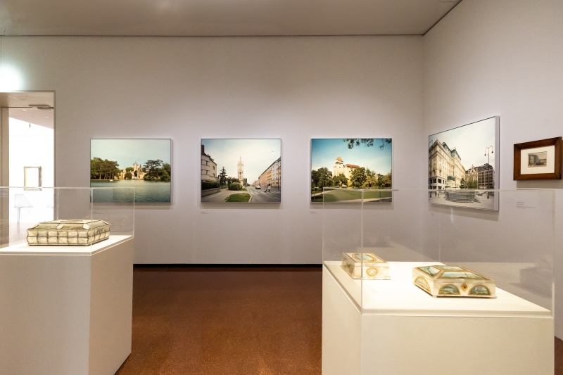 Das Foto zeigt einen hellen Ausstellungsraum mit vielen Bildern an den Wänden und zwei Vitrinen in der Mitte. Zu sehen ist ein Querschnitt aus historischen und zeitgenössischen Stadtansichten von Wien.
An den Wänden hängen großformatige Fotografien von Stefan Olah, die Wiener Plätze, Straßen und Gebäude zeigen. Ganz rechts ist ein Aquarell von Rudolf von Alt.
In den beiden Vitrinen stehen Dosen mit Bemalungen von Balthasar Wigand. Jede Bemalung zeigt eine andere Wienansicht – zum Beispiel den Stephansdom oder die Donau.