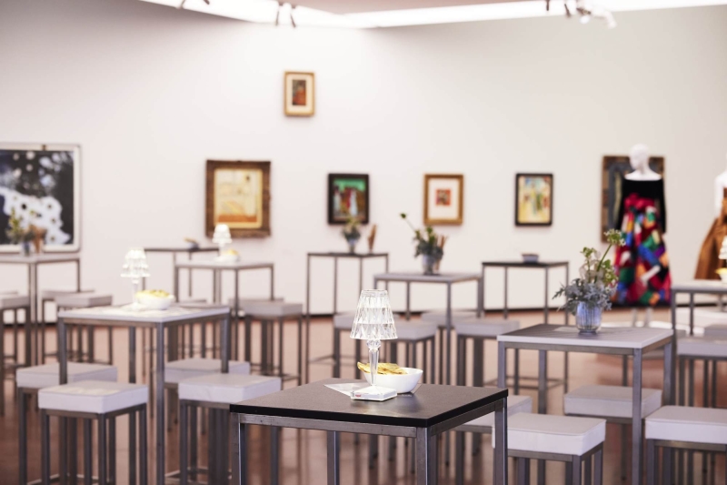 Moderne Galerie mit Stehtischen, Kunstwerken an Wänden und dekorativen Elementen wie Blumen und Lampen.