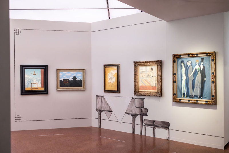 The photo shows part of the "Klimt⇄Warhol" exhibition in the Heidi Horten Collection museum in Vienna.
You can see a wall with several paintings. On the left are two surrealist paintings by René Magritte.
Below the paintings in the middle is an ornament in the shape of a grand piano. The decorations on the walls and the painted pieces of furniture were designed by the artist Markus Schinwald.