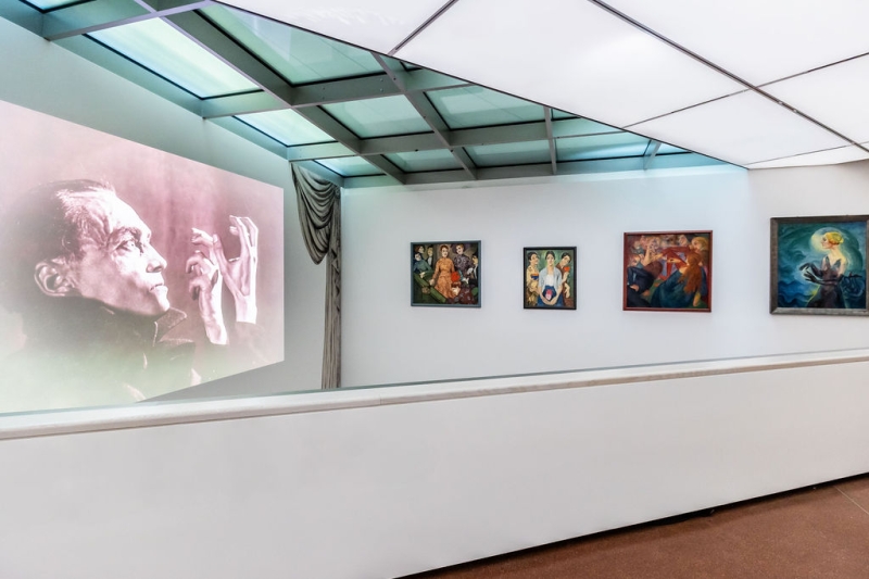 On the far left, you see a photo projected onto a wall.
It shows the actor Conrad Veidt in black and white.
He is raising his hands and looks serious.
To the right of it, there are four paintings:
– The first one on the left shows several people. In the center, a woman sits in an orange dress.
– The second one shows three women.
– The third one shows people from the story Decameron.
– The fourth is the painting “Terzetta”, with the woman raising her long, dark hands.
The colors in all the paintings are strong and bright.