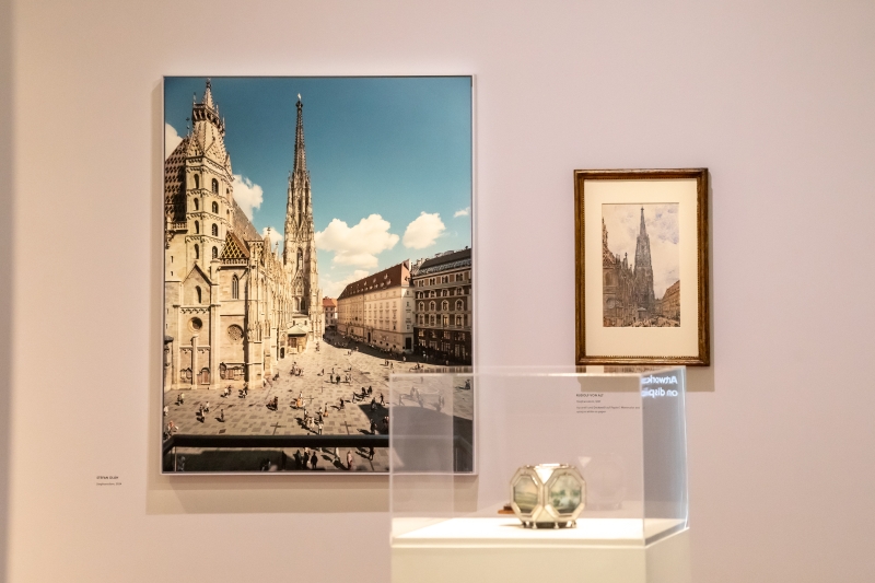 Auf dem Foto sieht man eine Ausstellungsansicht mit drei Kunstwerken, die sich mit der Stadt Wien beschäftigen.
Links hängt eine große Fotografie von Stefan Olah. Sie zeigt den Stephansdom bei schönem Wetter – mit blauem Himmel und Blick auf den belebten Platz davor.
Rechts daneben hängt ein kleineres Aquarell von Rudolf von Alt. Auch hier ist der Stephansdom zu sehen – fein gemalt, mit warmen Farben und vielen Details.
In der Vitrine darunter steht eine Wigand-Dose. Sie zeigt verschiedene Ansichten von Wien, unter anderem vom Stephansdom.