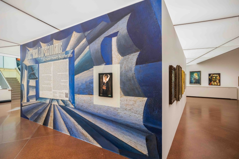 Exhibition entrance featuring a blue abstract wall, text panel, and framed artwork in a modern gallery setting.