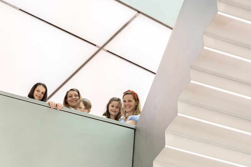 Auf dem Foto sieht man fünf Personen, darunter drei Kinder und zwei Erwachsene. Sie stehen auf einer Ausstellungsebene und schauen freundlich in die Kamera. Alle lächeln.
Rechts im Bild sieht man eine moderne, helle Treppe. Hinter den Personen sieht man die weiße Lichterdecke.