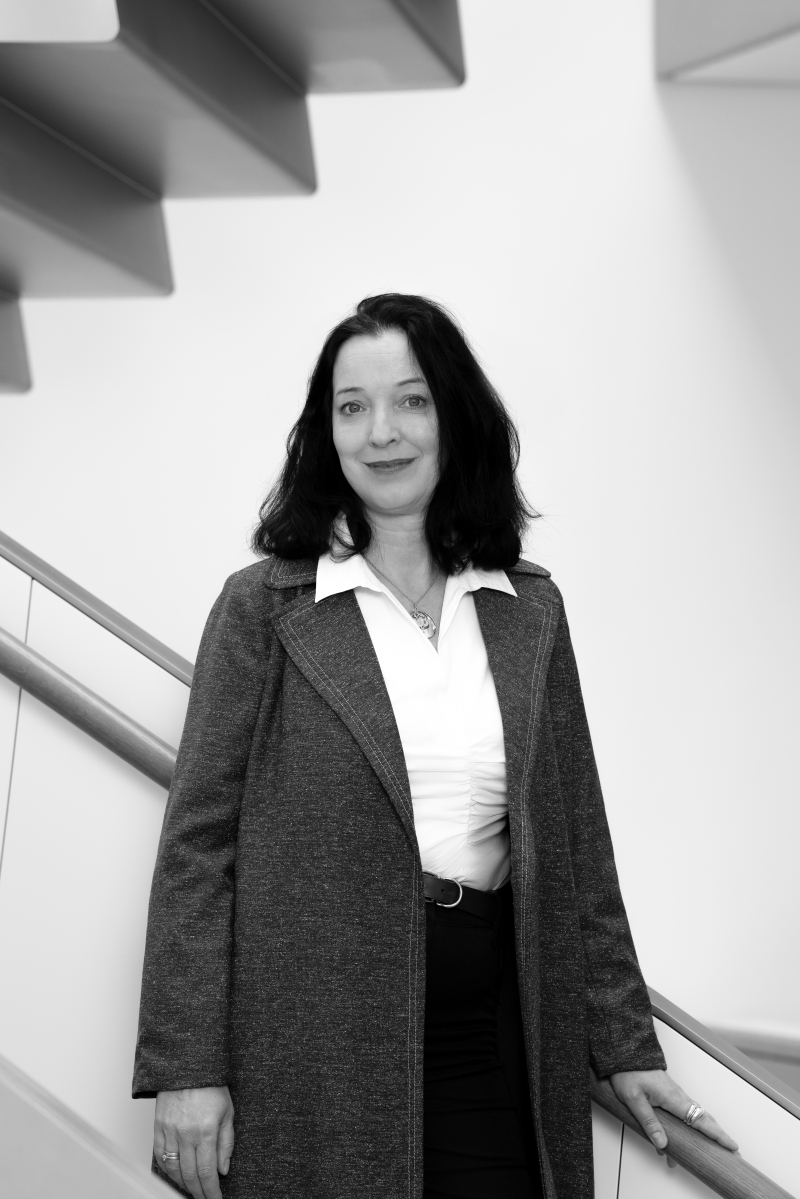 Auf dem Bild sieht man Susanne Wögerbauer.
Sie steht vor einer modernen Treppe mit klaren Linien.
Susanne trägt eine weiße Bluse, eine dunkle Hose und darüber einen langen, offenen Mantel in Grautönen.
Sie hat dunkles, schulterlanges Haar und blickt freundlich und selbstbewusst in die Kamera.
Das Foto ist in Schwarz-Weiß aufgenommen.