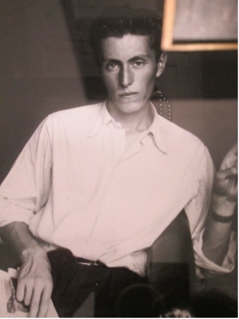 A young man with slicked-back hair sits casually, wearing a white shirt, with a contemplative expression and a hand resting on his knee.