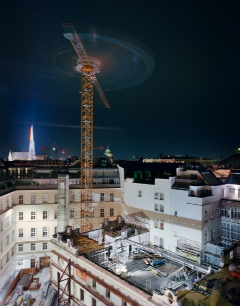 Baukran über einer Baustelle bei Nacht, umgeben von urbaner Architektur und beleuchteten Gebäuden.