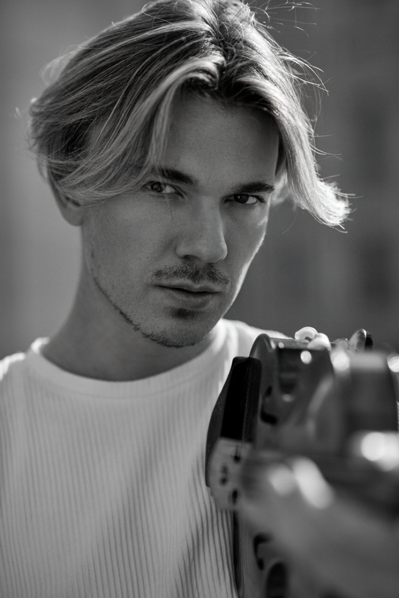 Junger Mann mit blonden, langen Haaren, trägt ein weißes Shirt und hält ein Instrument in der Hand, Blick direkt in die Kamera.