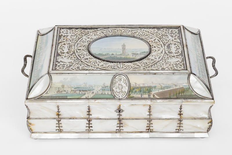Diese Schatulle wurde um das Jahr 1825 gemacht.
Die Aquarelle auf der Schatulle wurden von Balthasar Wigand gemalt.
Die Schatulle ist aus Perlmutt und Silber.
Sie sieht edel und kostbar aus.
An den Seiten hat sie kleine Henkel, mit denen man sie tragen kann.
Auf dem Deckel sieht man in der Mitte ein kleines Bild:
Es zeigt die Spinnerin am Kreuz – ein bekannter Ort in Wien.
Im Hintergrund sieht man die Stadt Wien und Menschen auf Pferden.
Rund um das Bild ist ein feines Muster aus Metall, mit Blättern und Figuren.
Auch an den Seiten der Schatulle sind gemalte Szenen von Wien zu sehen:
nämlich das äußere Burgtor mit Heldenplatz.