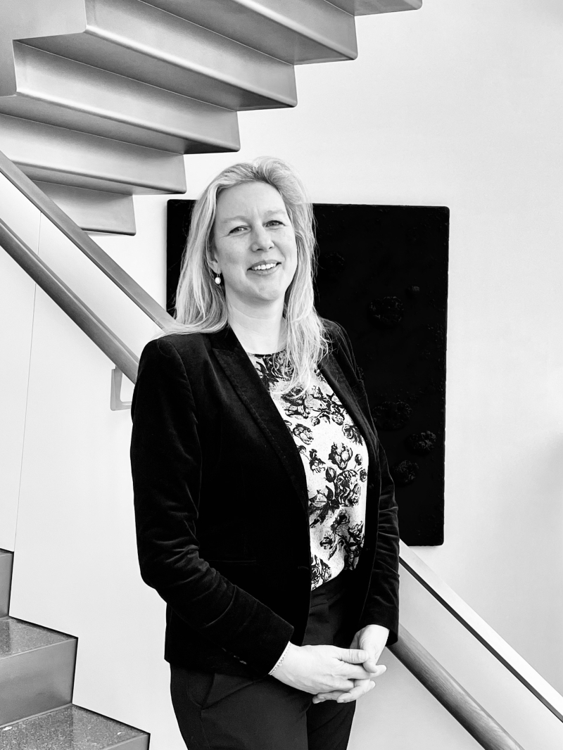 Auf dem Bild sieht man Anne-Sophie van Leeuwen.
Sie steht auf einer modernen Treppe in der Heidi Horten Collection.
Anne-Sophie trägt einen dunklen Blazer über einer gemusterten Bluse mit floralen Motiven.
Ihr Outfit wirkt klassisch und professionell.
Sie hat langes, offenes, helles Haar und blickt freundlich und gelassen in die Kamera.
Ihre Hände sind locker vor dem Körper gefaltet.
Im Hintergrund hängt ein Kunstwerk von Yves Klein an der Wand.
Das Foto ist in Schwarz-Weiß aufgenommen.