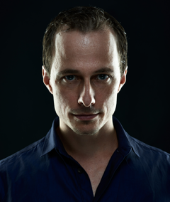 Mann mit kurzem Haar und ernstem Gesichtsausdruck, trägt ein dunkles Hemd, vor schwarzem Hintergrund.