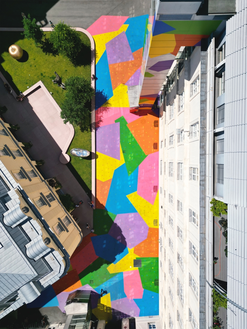 Auf dem Foto sieht man den Hanuschhof in Wien von oben. Die Häuser im Hof sind hell und der Boden und Lift sind in starken Farben bemalt – man erkennt große Flächen in Blau, Gelb, Grün, Pink, Schwarz und Lila.
In jeder Farbfläche sind viele kleine, bunte Punkte zu sehen. Diese Punkte zeigen, wie viel CO₂ in einem bestimmten Jahr ausgestoßen wurde – ein Punkt steht für eine Milliarde Tonnen.
Die Farben sind kräftig und leuchten im Sonnenlicht. Die Bemalung macht den Hof freundlich, auffällig und lebendig – und sie hat einen Zweck:
Die Spezialfarbe hilft dabei, die Hitze zu reduzieren, weil sie das Sonnenlicht besser zurückwirft als normaler Beton.
Dieses Kunstwerk heißt „CO₂ntext“. Es verbindet Umweltthema, Kunst und Stadtraum – und lädt zum Nachdenken und Verweilen ein.
Links oben sieht man einen Skulpturengarten von oben.