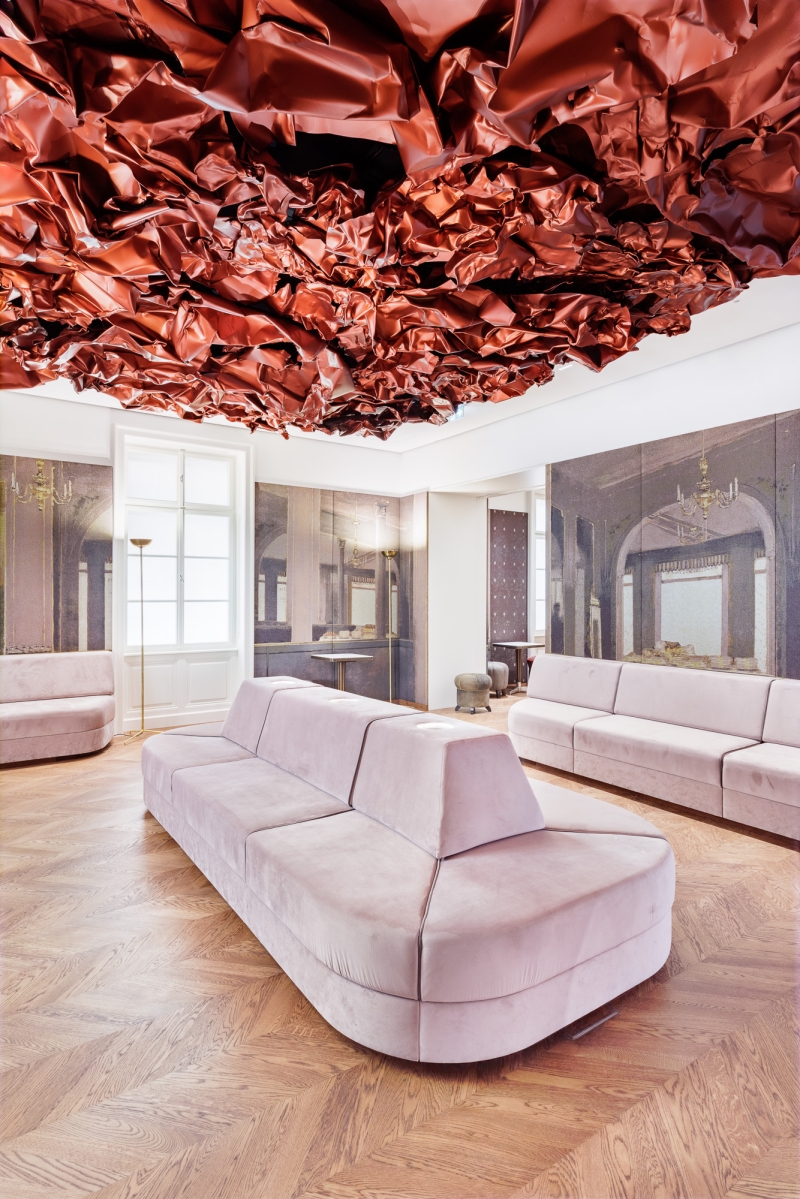The photo shows the Tea Room in the Heidi Horten Collection museum in Vienna.
A large, red work of art by Hans Kupelwieser hangs from the ceiling.
It looks like crumpled paper or shiny fabric, but
is made of aluminum.
There are lots of round windows on the back wall - they look like portholes.
Behind them are small, precious objects from Heidi Horten's collection.
This showcase wall was designed by Markus Schinwald.
The pink seating in the middle of the room is also part of his concept.
They invite you to sit down, look and relax.
The walls are covered with large fabric pictures,
reminiscent of an old café or castle room.