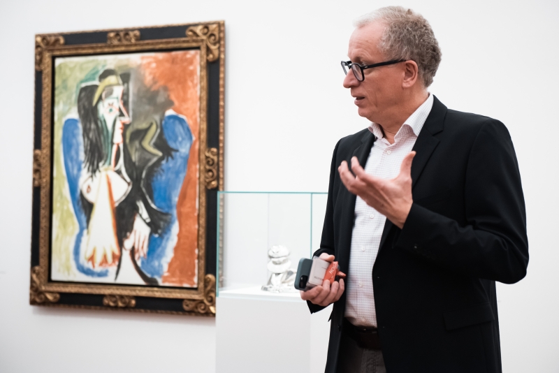 Mann in Anzug spricht vor einem Picasso-Gemälde und einer Glasvitrine mit einer Skulptur.