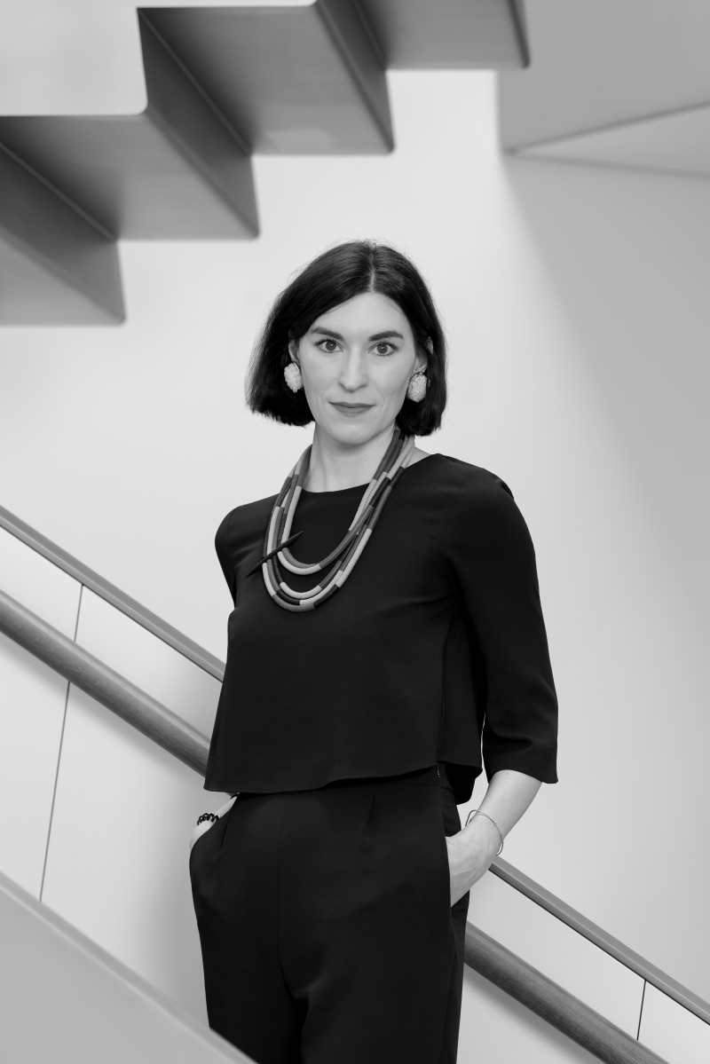 Auf dem Bild sieht man Pia Säf.
Sie steht vor einer modernen Treppe mit klaren Linien.
Pia trägt ein elegantes, dunkles Outfit mit weiter Dreiviertel-Ärmeln
und auffälligen Ketten und Ohrschmuck als Accessoires.
Sie hat dunkles, kinnlanges Haar und blickt ruhig und selbstbewusst in die Kamera.
Ihre Hände stecken locker in den Taschen, was ihr Auftreten souverän und entspannt wirken lässt.
Das Foto ist in Schwarz-Weiß aufgenommen.