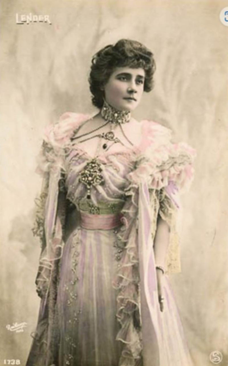 Mademoiselle Marcelle Lender poses in an elegant, pastel gown with ruffled sleeves and intricate jewelry against a soft backdrop.