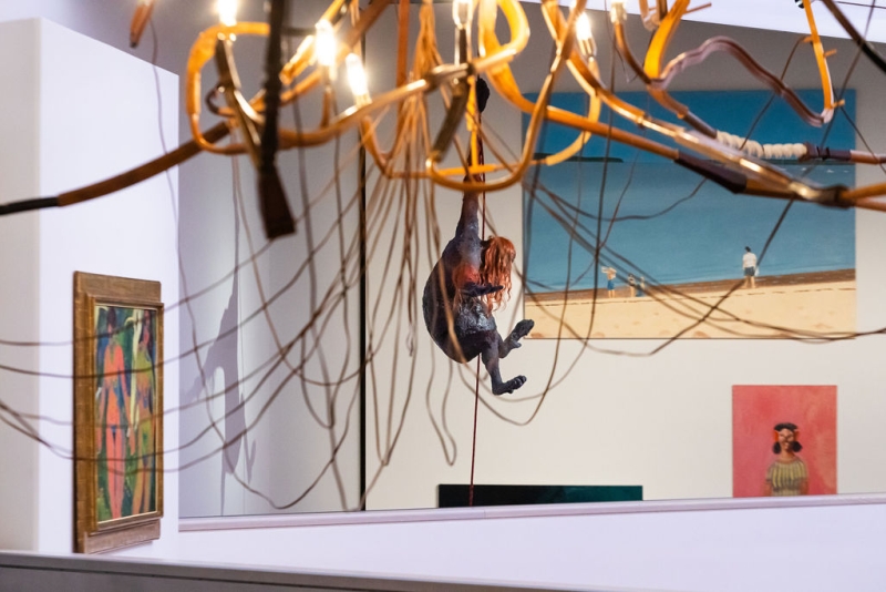 A figure hangs from a chandelier, surrounded by abstract art and paintings on the walls of a gallery.
