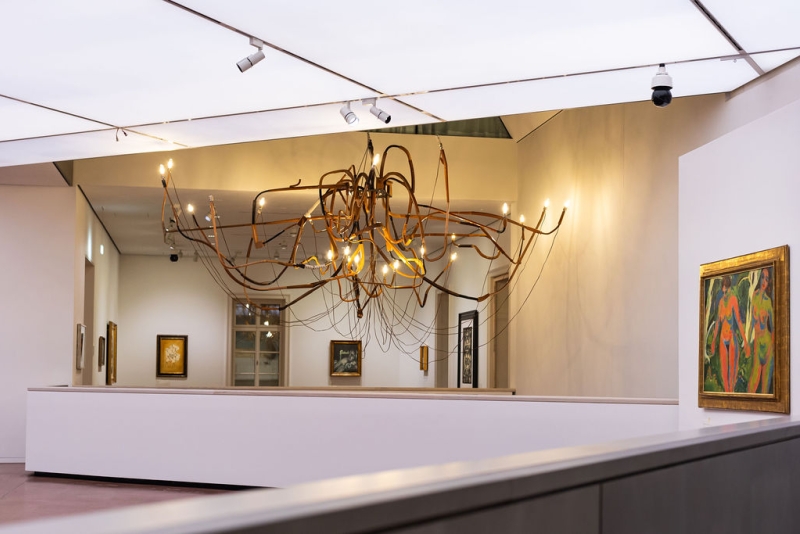 A modern chandelier with twisted branches and lights hangs in a gallery, featuring framed artworks on white walls.