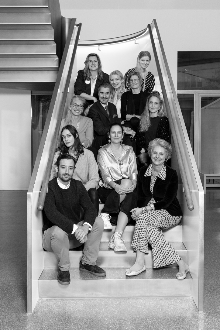 Auf dem Bild sieht man das Team der Kunstvermittlung in der Heidi Horten Collection. Die Gruppe sitzt und steht auf einer breiten, modernen Treppe im Inneren des Museums. Insgesamt sind dreizehn Personen auf dem Foto zu sehen – sie sitzen und stehen in mehreren Reihen, freundlich und offen der Kamera zugewandt. Alle tragen unterschiedliche Outfits, von leger bis elegant, was die Vielfalt und Individualität des Teams zeigt. Man sieht Vertrautheit und Zusammenhalt – das Team wirkt engagiert und gut gelaunt. Das Foto ist in Schwarz-Weiß aufgenommen.