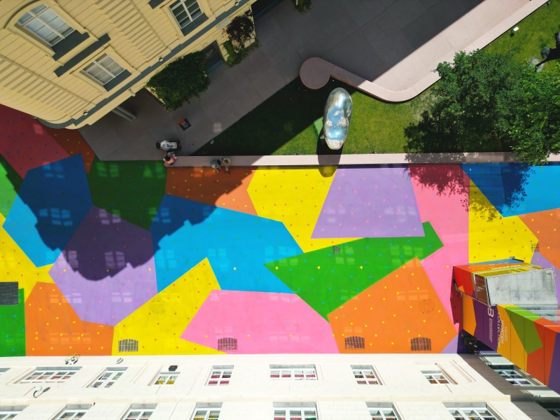 Bunte, geometrische Muster auf einem Platz, umgeben von Gebäuden und Grünflächen, mit Menschen und einer Skulptur.