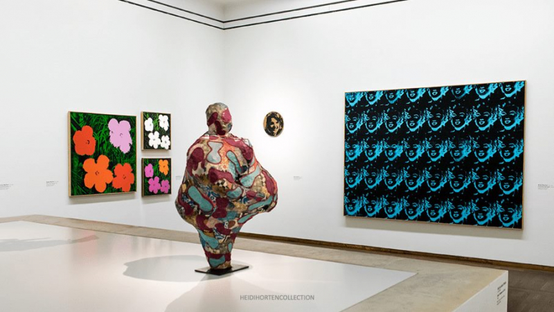 The photo shows a bright, modern exhibition space in the Leopold Museum. In the center is a large, colorful sculpture by Niki de Saint-Phalle. On the walls are works by Andy Warhol. His colorful flowers, a print with Mariylin Monroe and a small round work depicting Jacky Kennedy. The room is clear and calm, with a white floor and high ceilings.