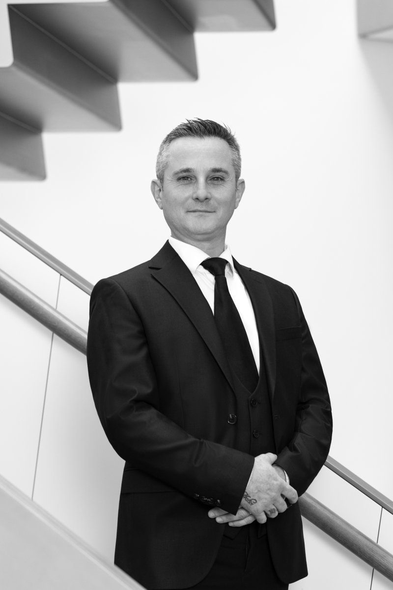 Auf dem Bild sieht man Shahin Ertekin. Er trägt einen dunklen Anzug, ein weißes Hemd und eine dunkle Krawatte. Er steht aufrecht und ruhig vor einer modernen Treppe mit klaren Linien. Seine Hände sind gefaltet, und er schaut freundlich und selbstbewusst in die Kamera. Das Foto ist in Schwarz-Weiß aufgenommen.