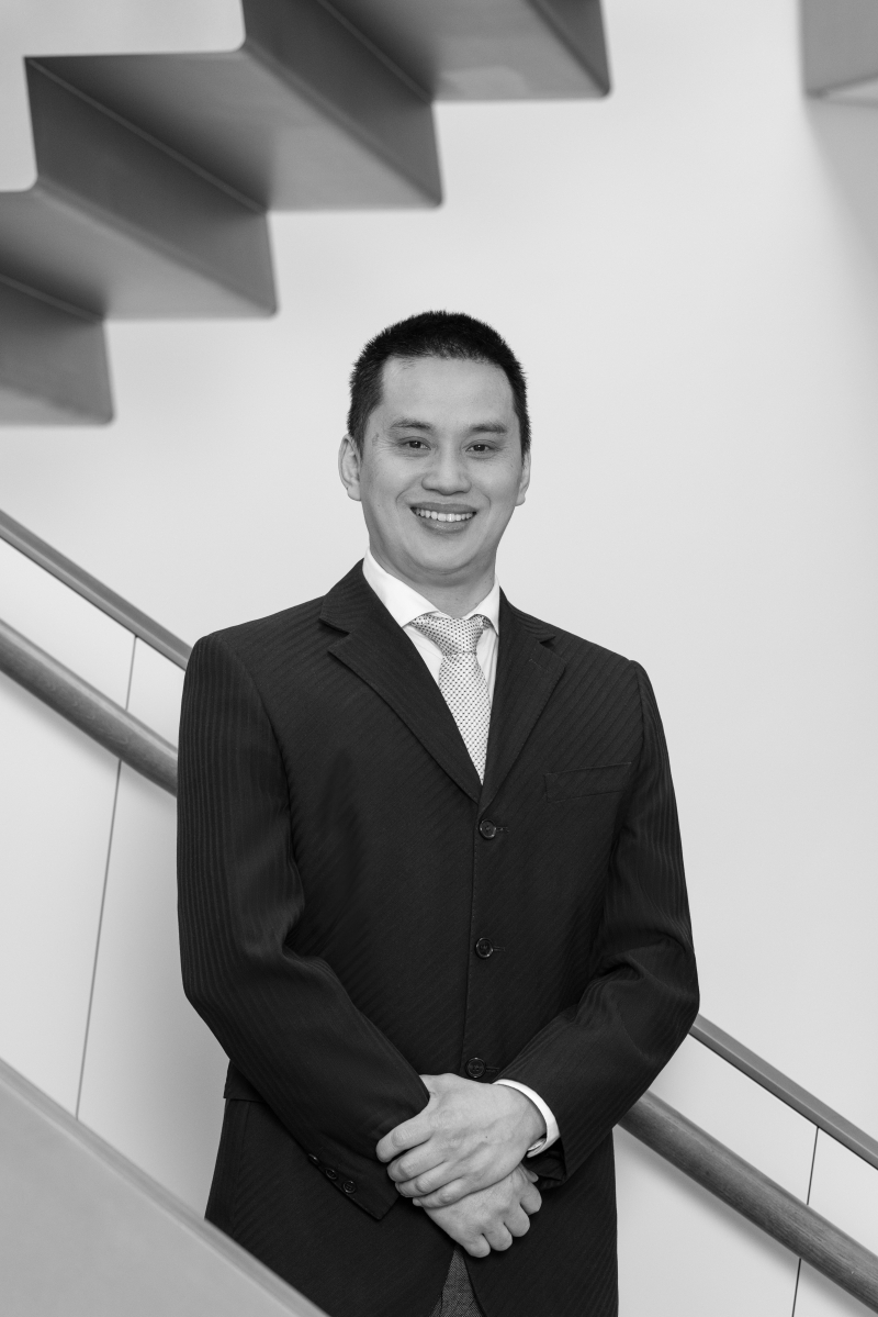 Auf dem Bild sieht man Min Ly.
Er trägt einen dunklen Anzug, ein weißes Hemd und eine dunkle Krawatte.
Er steht aufrecht und ruhig vor einer modernen Treppe mit klaren Linien.
Seine Hände sind gefaltet, und er schaut freundlich und selbstbewusst in die Kamera.
Das Foto ist in Schwarz-Weiß aufgenommen.