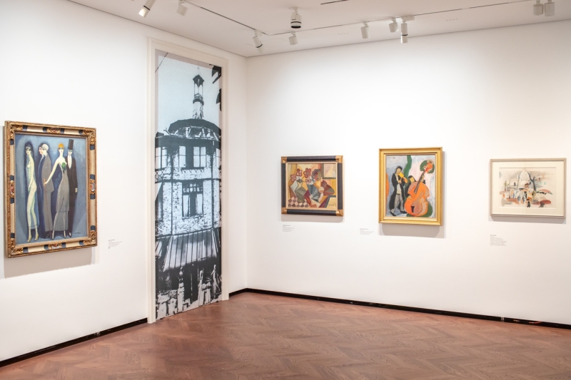 Ausstellungsraum mit mehreren Gemälden, darunter Figuren und eine Musikergruppe, sowie einer großen Wandgrafik.