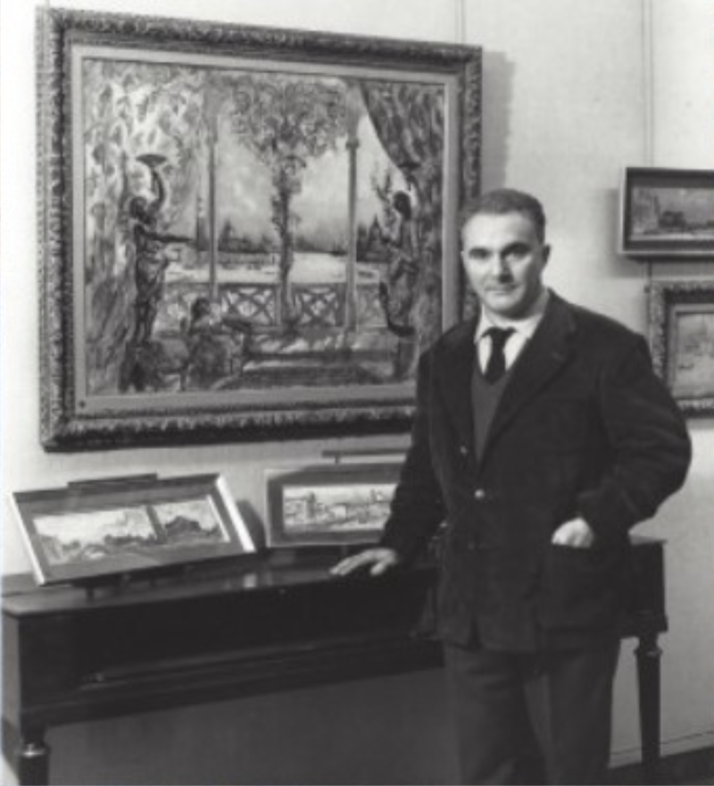Man in a dark coat stands beside a piano, with a large painting and smaller artworks displayed on the wall behind him.