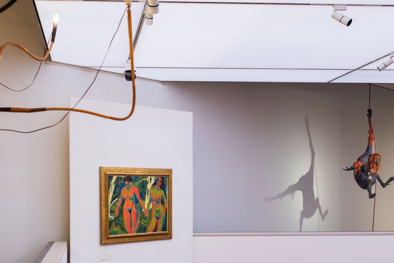 Colorful painting of two figures on a white wall, with a hanging sculpture casting a shadow in an art gallery.