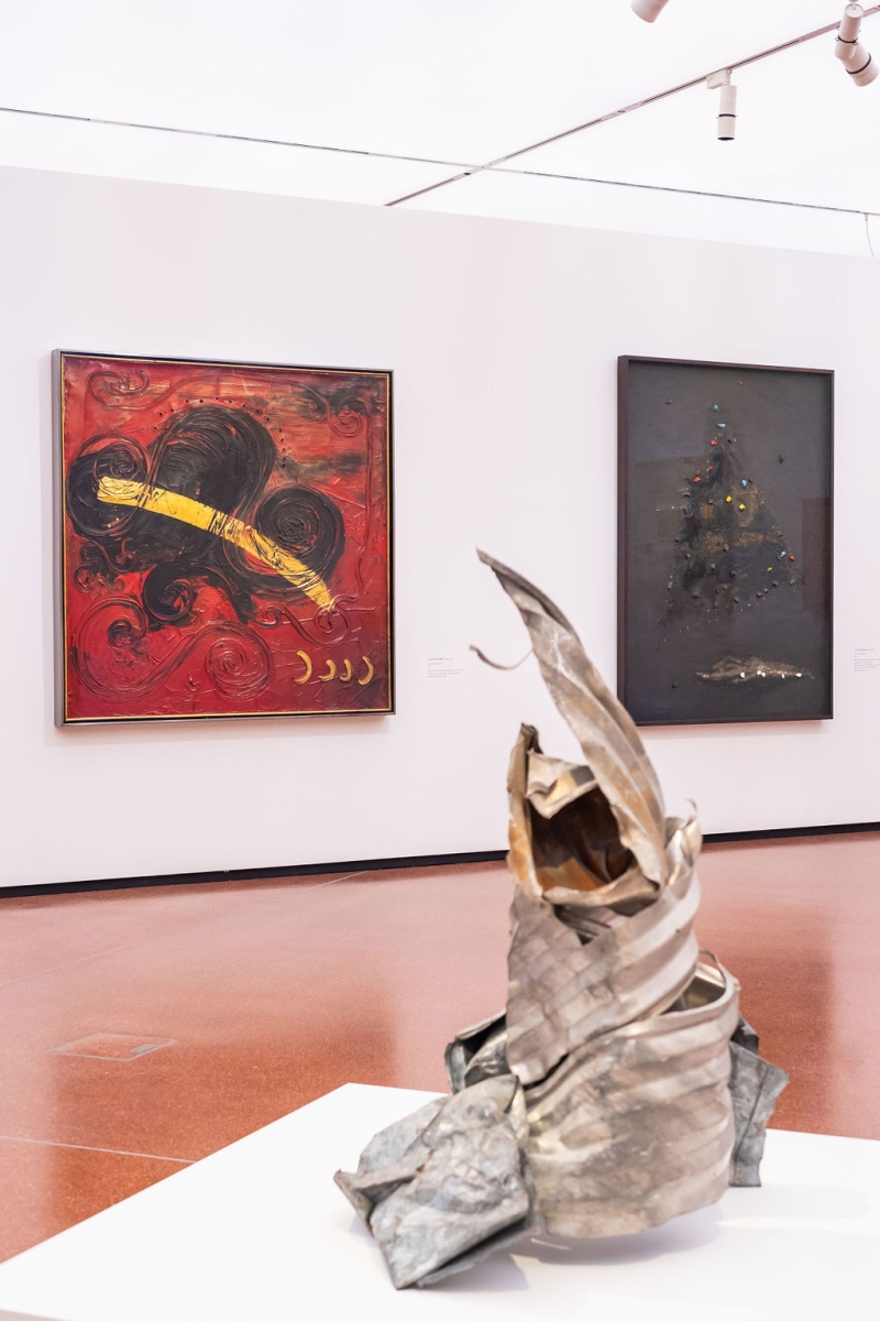 Abstract red painting with yellow streak, dark artwork on right, and a twisted metal sculpture in foreground.
